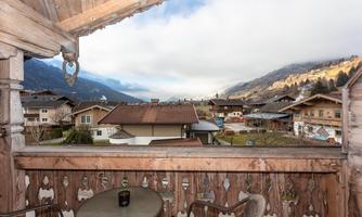 A balcony view with traditional wooden siding and a table. In the background, houses and mountains are visible under a partly cloudy sky.