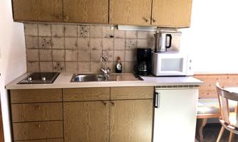 A small, bright kitchen with wood cabinets and a sink. On the countertop, there is a kettle and a microwave.