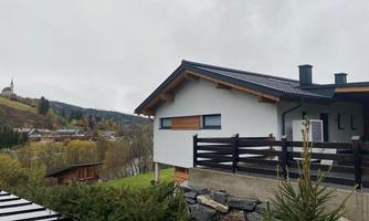 A modern house with a stylish roof and a wooden terrace. In the background, green meadows and wooded hills can be seen.