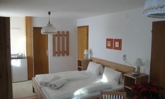 A bright room with a double bed and a small dining area. The walls are in neutral colors, and there is a small kitchenette in the background.