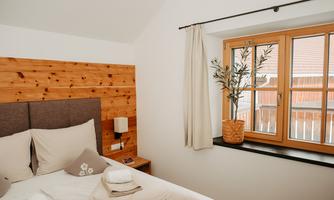 A cozy bedroom with a wooden headboard and a window that lets in plenty of light. On the nightstand, there is a small lamp and a plant in a basket.