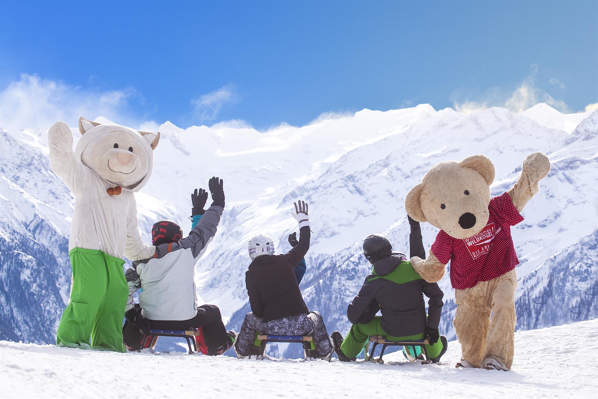 A group of people is sitting in the snow and waving. Two persons are wearing animal costumes and the mountains are visible in the background.