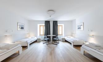 A bright, modern room with three single beds and a table. The walls are white, and there are large windows with curtains.