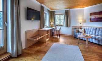 A bright and cozy room with wooden floors and modern furniture. There is a desk, a sofa, and large windows for plenty of daylight.