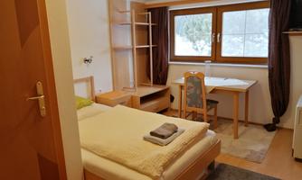 A simple bedroom with a single bed, a table, and a chair. Large windows provide plenty of daylight.