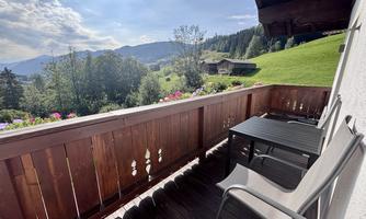 A balcony with two chairs and a table offers a view of a green landscape and mountains. The flowers on the railing add a cozy atmosphere.