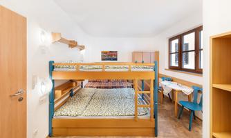 A bright room with a bunk bed and a small table. The walls are white, and there are large windows that let in a lot of light.