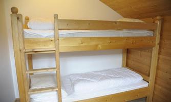 A simple wooden bunk bed stands in a bright room. There are mattresses with white bedcovers on the top and bottom.