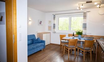 A cozy living area with a blue sofa and a dining table. Large windows provide plenty of natural light.