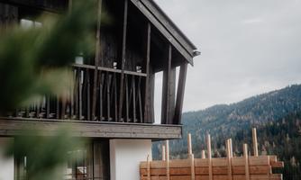 A modern wooden house with a balcony and a beautiful mountain landscape in the background. The clouds in the sky give the scene a calm atmosphere.