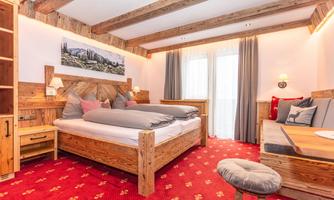 A cozy hotel room with wooden furniture and a red carpet. The room features a large bed, a seating area, and windows with curtains.