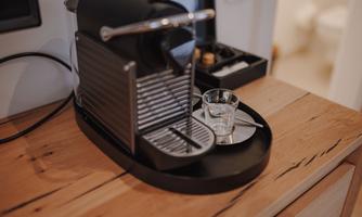 A modern coffee machine on a wooden board with a small glass. In the background, other kitchen utensils can be seen.