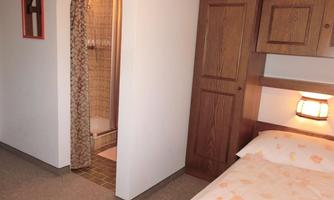 A simple room with a bed and an adjoining bathroom. The walls are light, and there is wooden furniture as well as a warm light source.