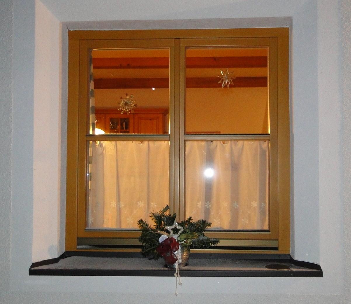 A window with a yellow frame, radiating warm light from an interior. On the windowsill, there is a decorative arrangement of pine branches and a red ribbon.
