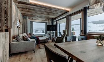 A modern living room with large windows and a view of a snowy landscape. Wood paneling and cozy furniture create an inviting atmosphere.