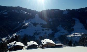 A picturesque winter landscape with snow-covered mountains and traditional wooden houses. The sun shines brightly in the clear sky.