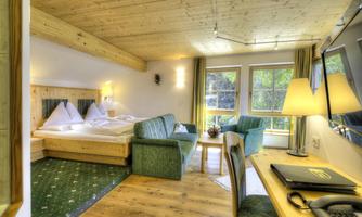A cozy hotel room with wood decor, a double bed, and a small sofa. Large windows offer a view of greenery and plenty of daylight.