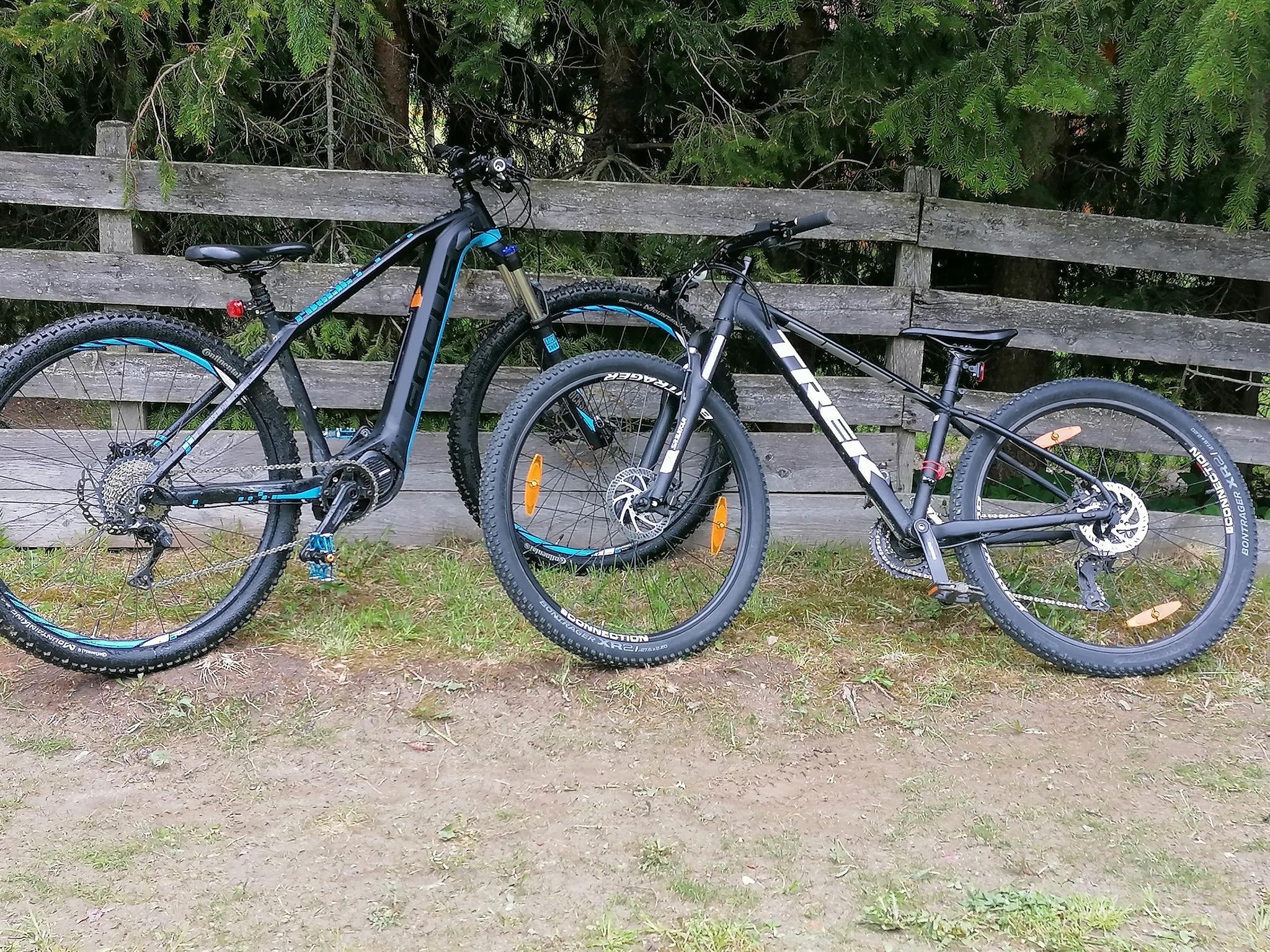Two bicycles are next to a wooden bench. In the background, trees can be seen.