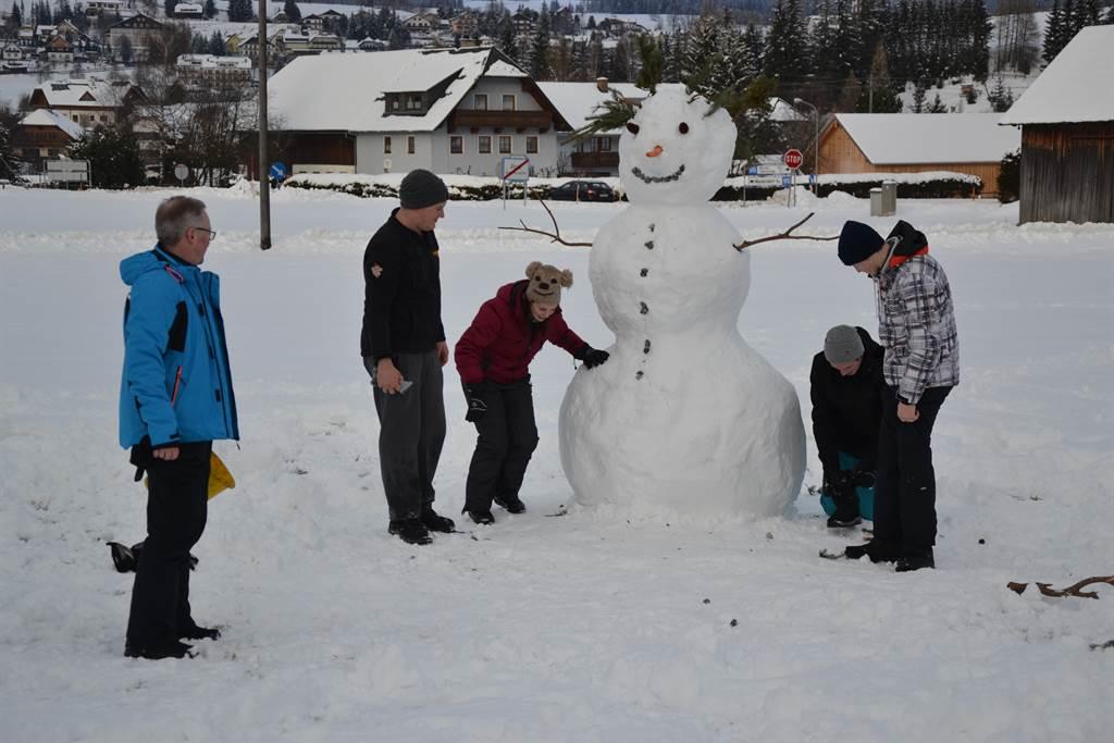 A group of people is building a large snowman in the snow. In the background, there are houses and a snow-covered landscape.