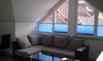A cozy living room with a large gray sofa and a glass table. The windows have blue blinds and offer a view of rooftops.