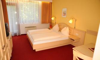 A cozy hotel room with a double bed, two pillows, and a large window front. The walls are painted in a warm yellow tone, and there is a small table as well as a TV.