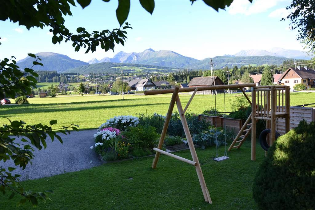 A beautiful garden with a play tower and a swing. In the background, green meadows and high mountains stretch under a blue sky.