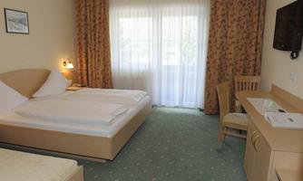 A bright hotel room with a double bed and a desk. Large windows allow plenty of daylight to enter and offer a view outside.