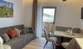 A modern and cozy living room with a couch and a dining table. Large windows offer a view of the mountains and the outdoor area.