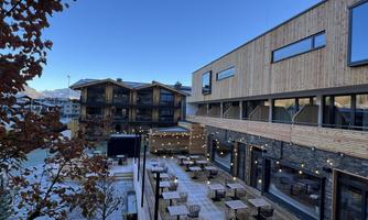 A modern building with a wooden facade and large windows. In the foreground, there is a cozy terrace with tables and chairs.