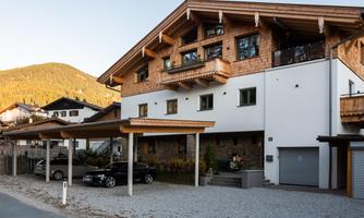 A modern house with wooden cladding and a balcony. In front of the house, there are covered parking spaces for cars.