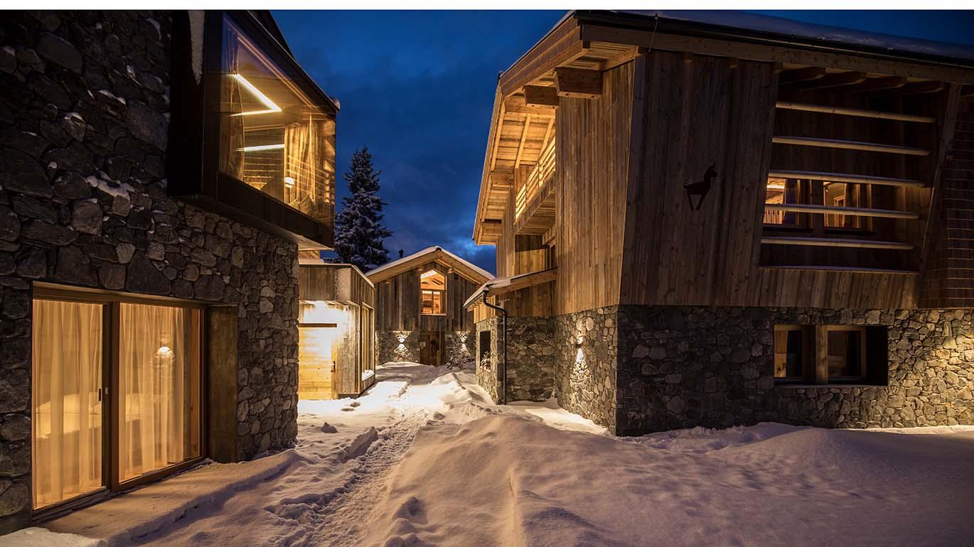 A wintry village scene with wooden chalets surrounded by snow. The warm lights in the windows give the scene an inviting atmosphere.