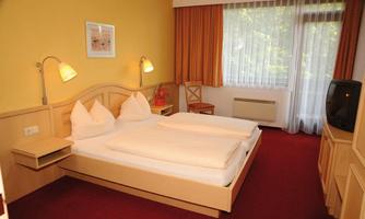 A cozy hotel room with two single beds and light wood decor. The walls are painted in warm yellow tones, and a large window lets in plenty of daylight.