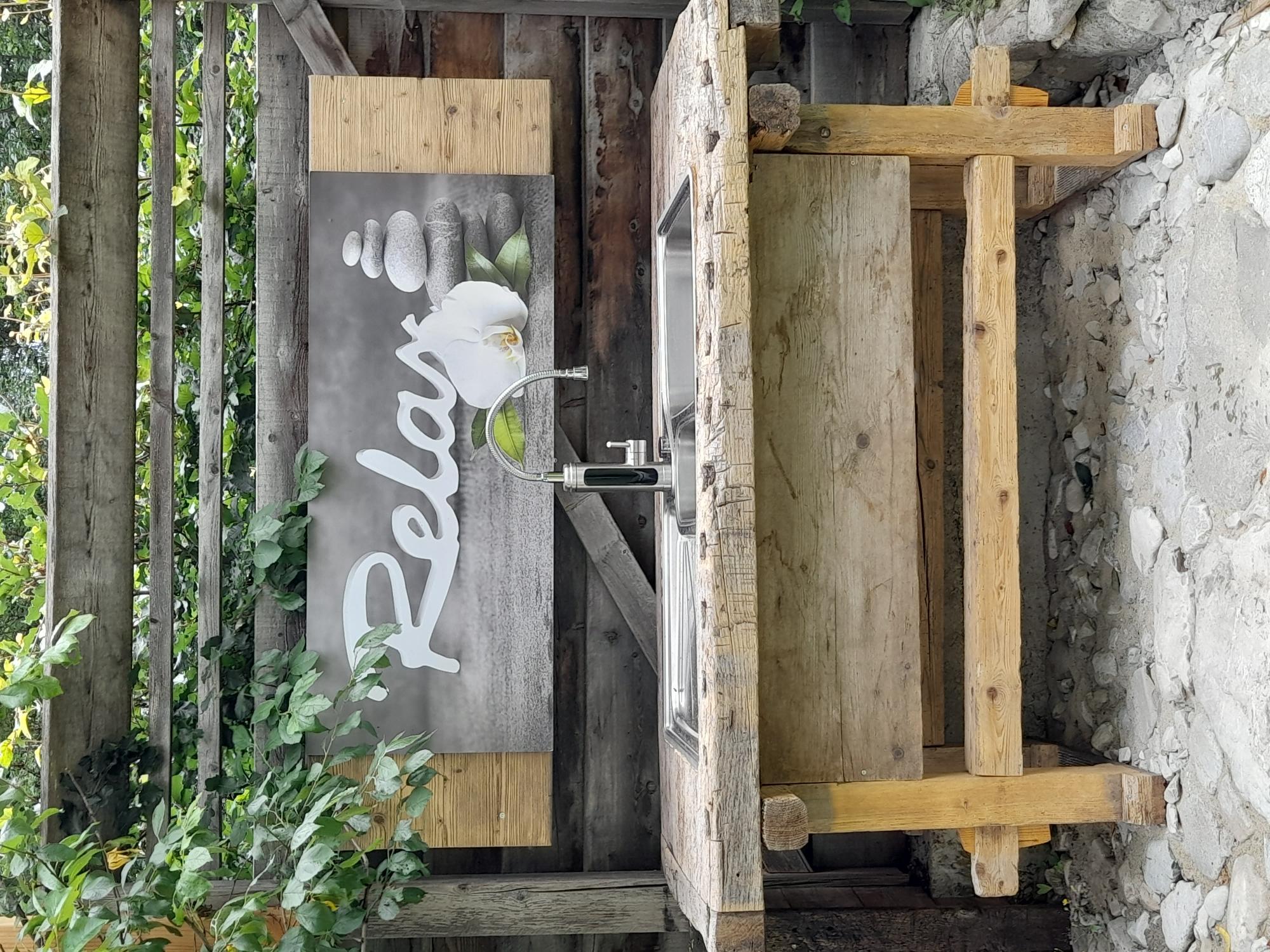 A rustic wooden washing station outdoors with a modern faucet. In the background, there is a relaxing "Relax" sign and some green plants.