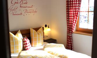 A cozy bedroom with yellow-checked bed linen and colorful cushions. A decorative saying in German script hangs on the wall.