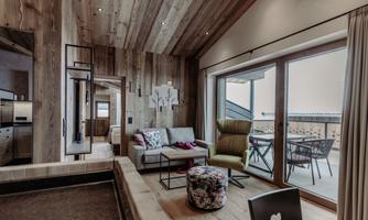 A modern living room with wooden paneling and large windows. A cozy sofa and a beautiful view of the balcony are available.