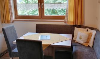 A cozy dining area with a round table and a corner bench. The windows are decorated with yellow curtains and let in plenty of light.