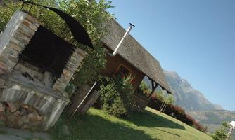 A stone barbecue area is in the foreground. In the background, a wooden house can be seen against a mountainous landscape.