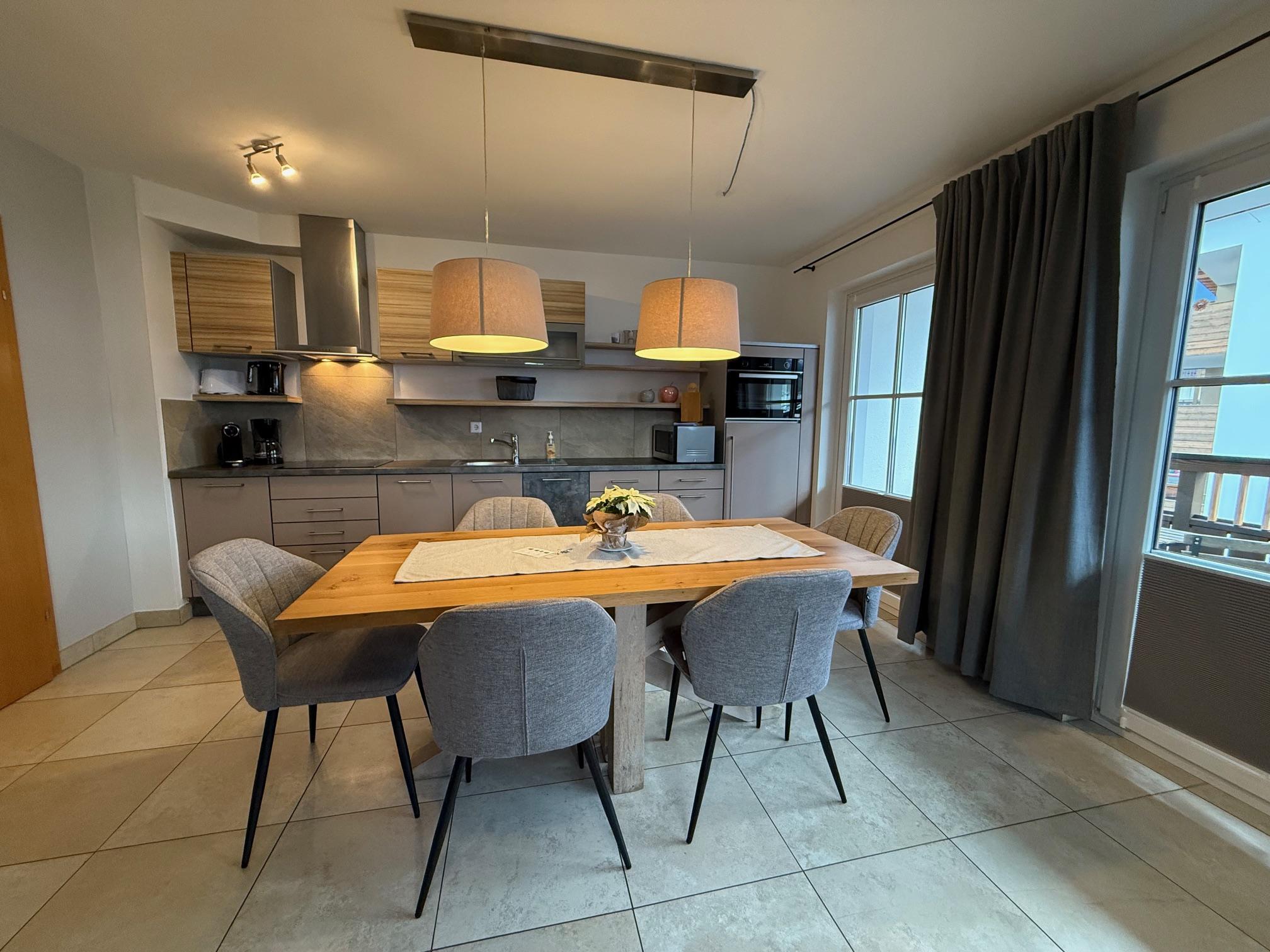 A modern kitchen with a large wooden table and comfortable chairs. The space is bright and welcoming, with windows and minimalist decoration.