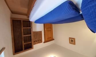 A simple bedroom with a double bed and blue pillows. There is a wooden wardrobe and a warm, inviting atmosphere.