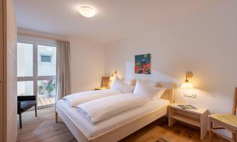 A bright bedroom with a double bed and wooden floor. There is a chair, a small table, and a window with a view outside.