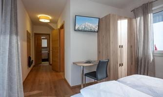 A modern room with a double bed and a table. A television hangs on the wall, and there is a view of a hallway with wooden details.