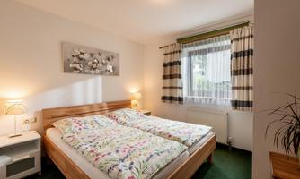 A cozy bedroom with a double bed and colorful bedcovers. The room has large windows and friendly furniture.