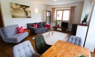 A bright living room with modern furniture and a large window. There is a cozy sofa, a wooden dining table, and a TV corner.