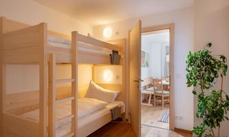 A cozy room with a bunk bed and warm wood. In the background, a living area with a dining table and plants is visible.