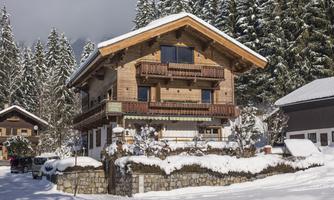 A cozy wooden house in a snowy landscape. In the background, tall green trees can be seen.