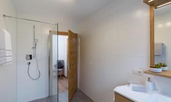 A modern bathroom with a glass shower and a wooden vanity. The walls are bright and neutral, and there is a view of an adjacent room.