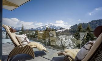 A beautiful balcony with comfortable loungers and a breathtaking view of snow-capped mountains. The sky is clear and blue, surrounded by green trees.
