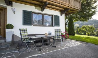 A cozy outdoor area with a table and chairs in front of a charming house. In the background, you can see a green meadow and trees.