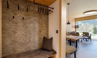 A modern entrance area with a bench and simple coat hooks. In the background, a bright living area with a view of nature can be seen.
