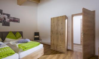 A modern bedroom with a double bed, green pillows, and a wooden wardrobe. A door leads into an adjacent room.